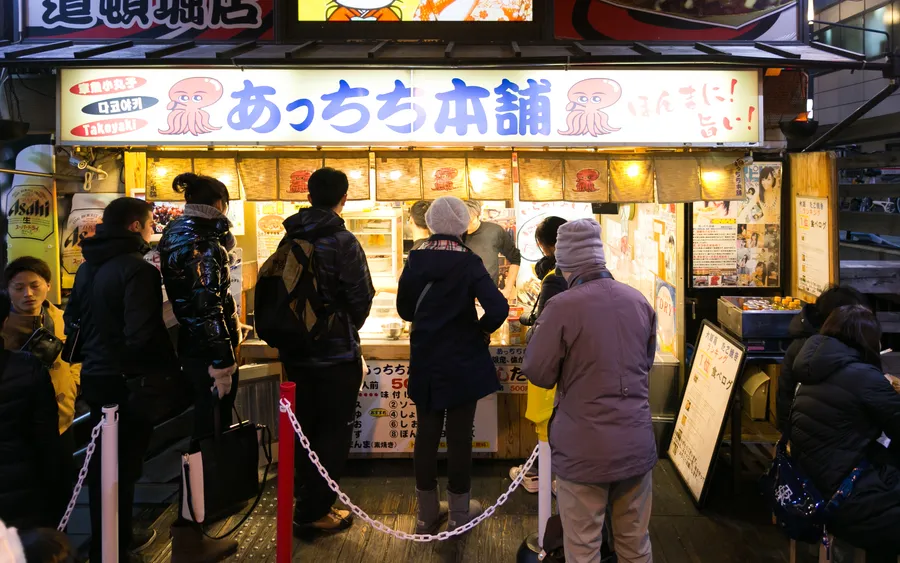 A takoyaki shop storefront with a queue of customers in Dotonbori, Osaka